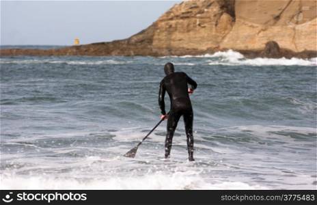 One man paddles out into the ocean on long paddle board near rock formation