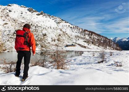 One man young male hiker stands in front of a alpine lake looking to the snowy landscape, rear view. Red jacket, black pants. Large copy-space at top-right. Buffet Lake, Champorcher, Val d&rsquo;aosta, Italy, Europe.