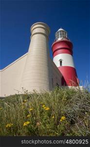 One of the two buttraces of the Cape Agulhas lighthouse stand prominent in front of the lighhouse itself as viewed from a low side angle.