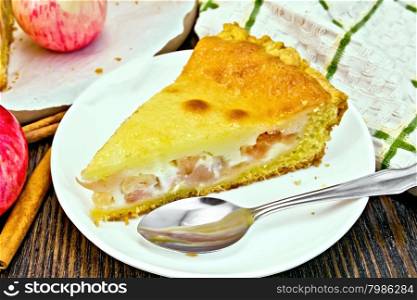 One slice of apple pie with cream sauce in white plate, spoon, cinnamon, apples and a napkin on a dark wooden board