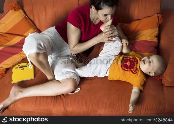 One year old baby and his mother are playing together on a couch. The mother is kissing his little foot.It is a typical home enviroment.