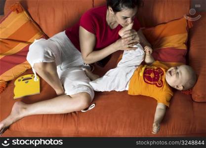 One year old baby and his mother are playing together on a couch. The mother is kissing his little foot.It is a typical home enviroment.