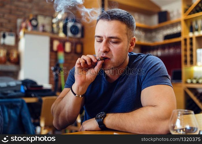 One Young adult caucasian man sitting alone by the table at home or restaurant holding the cigar smoking wearing t-shirt in day looking down smoke around his head