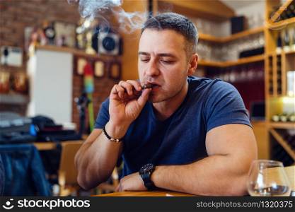 One Young adult caucasian man sitting alone by the table at home or restaurant holding the cigar smoking wearing t-shirt in day looking down smoke around his head