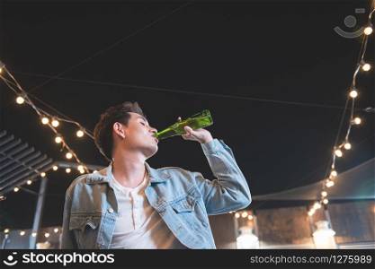 One young asian man drink bottle of beer. Side view of handsome hipster men hand holding beer bottle and drink while sitting at the bar counter on rooftop floor of nightclub party at restaurant.