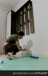 One young beautiful Caucasian brunette girl, squatting, paints a wall with white paint with a brush during the day in the living room, close-up side view.. A young girl paints a wall with a brush.