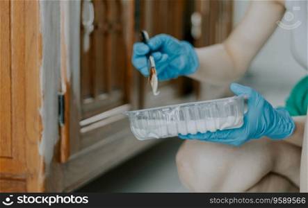 One young Caucasian unrecognizable girl in blue disposable gloves holds a transparent box of white paint and with a narrow brush for painting the cabinet, sitting in the room during the day, close-up side view.. A young girl paints an old wardrobe.