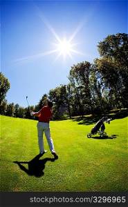 One young white male golfer, red shirt and white pants, goes for the green on the fairway. Vertical frame, clear summer day, back-light.
