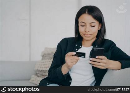 Online banking. Female holding credit card and smartphone, makes easy fast secure money transfer, using mobile bank app at home. Modern young woman makes purchases on internet, sitting on couch.. Online banking. Female holds credit card, phone, makes money transfer using mobile bank app at home