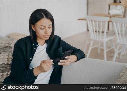 Online banking. Focused female tenant holds mobile phone credit bank card pay rental, makes purchases. Woman does easy fast secure money transfer in e-bank application sitting on couch at home.. Online banking. Female tenant hold credit bank card pay rental by mobile phone app sitting on couch
