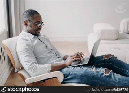 Online freelance job. Handsome african american man in front of pc. Relaxed happy businessman in formal wear is sitting in cosy chair at his apartment. Startup project, remote work.. Online freelance job. Handsome african american man in cosy chair in front of pc at his apartment.