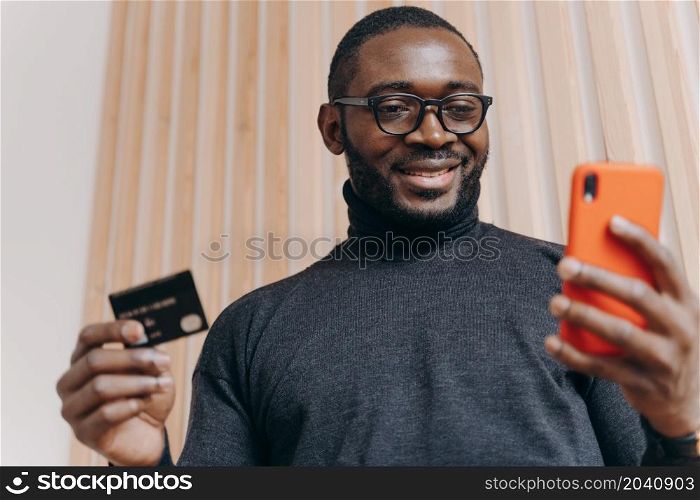 Online payment. Cheerful African american businessman holding credit card and smartphone for shopping in internet, with happy smile looking on phone screen satisfied with successful transaction. Cheerful African american businessman holding credit card and smartphone for shopping in internet