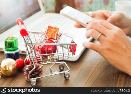 Online payment, Close up at gift boxes in shopping cart and Christmas decoration, woman hands using smartphone and laptop computer in background