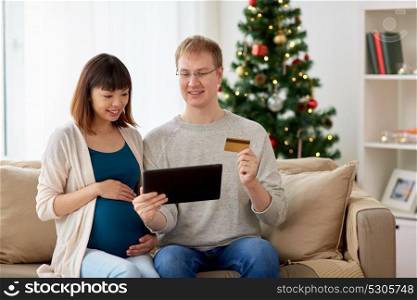online shopping, winter holidays and people concept - happy husband and his pregnant wife with tablet pc computer and credit card home at christmas. man and pregnant wife shopping online at christmas