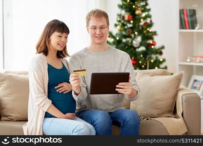 online shopping, winter holidays and people concept - happy husband and his pregnant wife with tablet pc computer and credit card home at christmas. man and pregnant wife shopping online at christmas