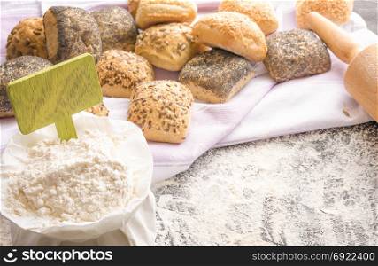 Open bag of flour with a green wooden banner stuck in it surrounded by freshly baked bread rolls decorated with seeds, displayed on a towel.
