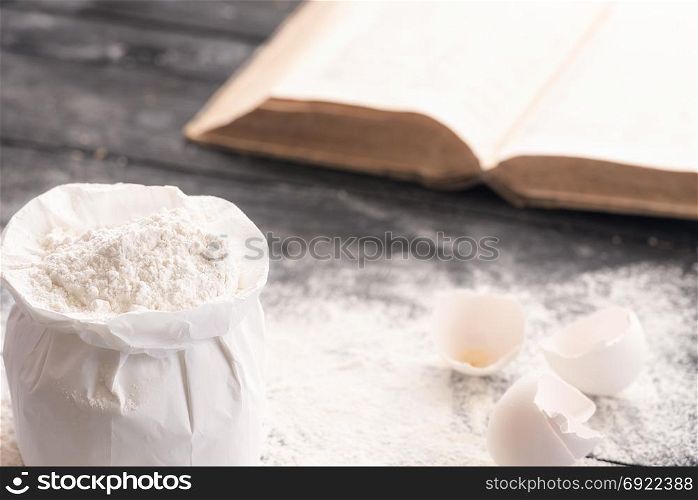 Open bag of wheat flour on a rustic wooden table covered with flour, egg shells and an open recipe book in the background.