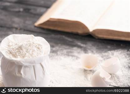 Open bag of wheat flour on a rustic wooden table covered with flour, egg shells and an open recipe book in the background.