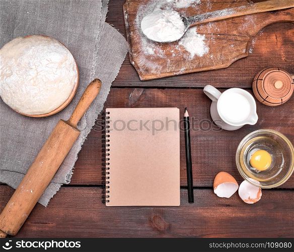 open blank notebook and black wooden pencil in the middle of the dough and ingredients for cooking, top view