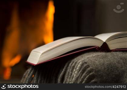 Open book by fireplace. Peaceful closeup of open book reosting n a arm rest of a couch. Warm fireplace on background.