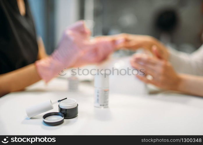 Open bottle with white nail polish and brush, manicure master in pink gloves applying varnish to female client on background, beauty salon. Manicurist doing fingernail care cosmetic procedure