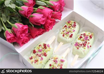open box with cakes in the form of popsicle on a stick in light green glaze and flower and pistachio topping. and a large bouquet of bright pink roses.