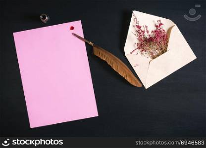 Open envelope full with red flowers, an antique feather pen on a pink paper note with a red ink splash on a black wooden background.