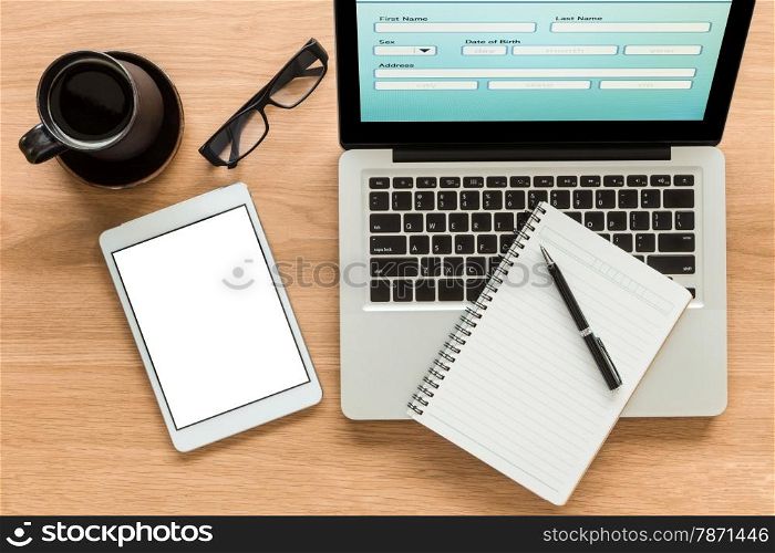 Open laptop shows blank information form and digital tablet with isolate screen on wooden table. Blank diary book and pen on laptop, glasses and a cup of coffee on workspace. Top view image for mock up concept.