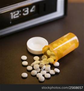 Open medicine bottle with pills spilling out onto table with clock in background.