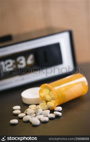 Open medicine bottle with pills spilling out onto table with clock in background.