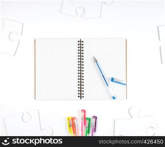 open notebook in a cell and a blue pen on a white background, next to large blank puzzles, top view