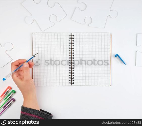 open notebook in a cell, hand holding a blue pen, next to it there are big white puzzles, top view