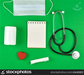 open notebook in line with white sheets, medical mask, stethoscope and bandage on a green background