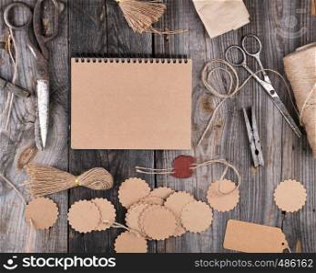 open notepad with brown empty sheets on a gray wooden table, next to it are a rope, paper tags and scissors, top view