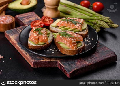 Open sandwich or toast. Grain bread with salmon, avocado and sesame seeds. Healthy snack, fat and omega 3 source. Toast sandwich with butter, avocado and salmon, decorated with arugula and sesame seeds, on a black stone background