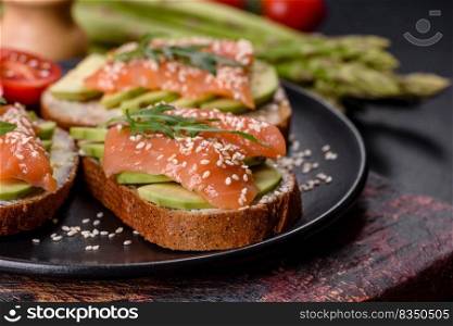 Open sandwich or toast. Grain bread with salmon, avocado and sesame seeds. Healthy snack, fat and omega 3 source. Toast sandwich with butter, avocado and salmon, decorated with arugula and sesame seeds, on a black stone background