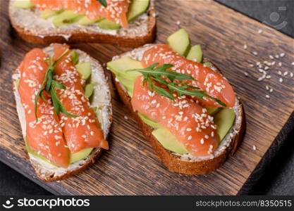 Open sandwich or toast. Grain bread with salmon, avocado and sesame seeds. Hea<hy snack, fat andω3 source. Toast sandwich with butter, avocado and salmon, decorated with arugula and sesame seeds, on a black sto≠background