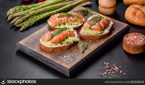 Open sandwich or toast. Grain bread with salmon, avocado and sesame seeds. Healthy snack, fat and omega 3 source. Toast sandwich with butter, avocado and salmon, decorated with arugula and sesame seeds, on a black stone background