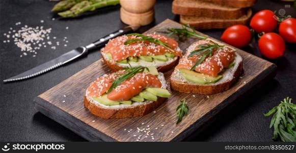 Open sandwich or toast. Grain bread with salmon, avocado and sesame seeds. Healthy snack, fat and omega 3 source. Toast sandwich with butter, avocado and salmon, decorated with arugula and sesame seeds, on a black stone background