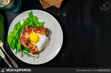 Open sandwich with bacon, fried egg and celery leaves on a slice of rye sourdough bread with pumpkin seeds on a white plate. Healthy food concept, traditional craft bread, top view with copy space
