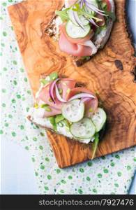 Open sandwiches with ham, cucumber, arugula and soft cheese on a crusty slice of rye bread over olive wood board, top view