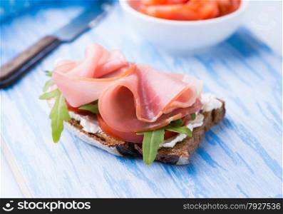 Open sandwiches with ham, tomato and arugula on blue background, selective focus