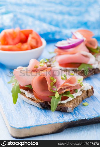 Open sandwiches with ham, tomato and arugula over blue background, selective focus