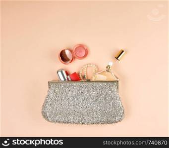 open silver clutch and cosmetics fell out of the middle, peach background, top view, copy space