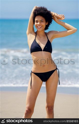 Optimistic African American female in bikini looking at camera with hand behind head while standing on sandy coast near sea. Delighted black woman on seashore