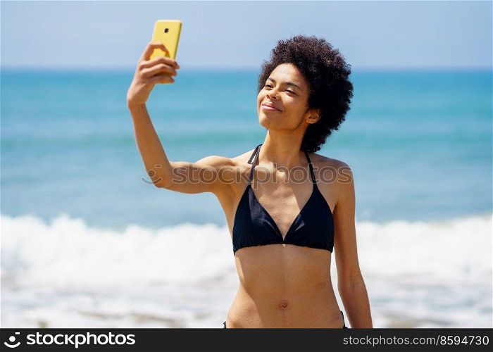 Optimistic African American female tourist in swimwear taking self portrait on mobile phone while standing near waving sea during summer vacation. Content black woman in bikini taking selfie on beach