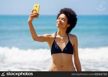 Optimistic African American female tourist in swimwear taking self portrait on mobile phone while standing near waving sea during summer vacation. Content black woman in bikini taking selfie on beach