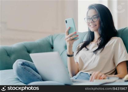 Optimistic brunette in casual clothes and glasses, focused on smartphone, shops online, watches training webinar on sofa.