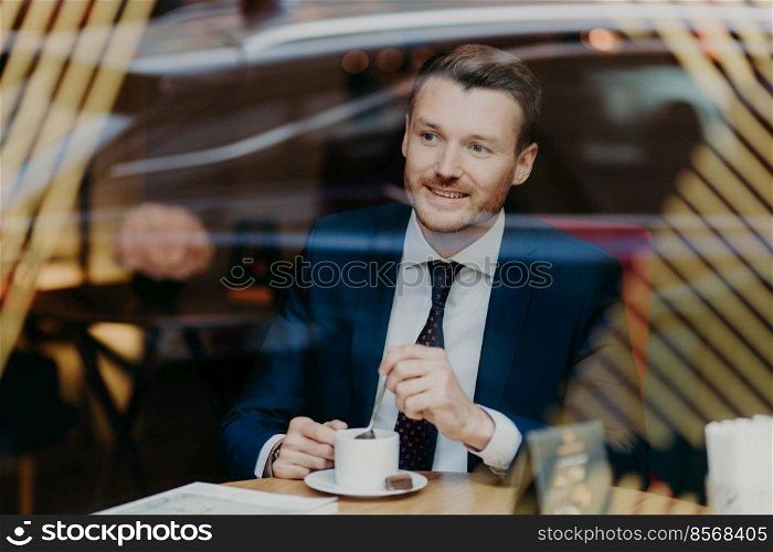 Optimistic cheerful young businessman has thoughful expression, dressed in formal clothes, looks through window in cafeteria, satisfied with results of work. People, leisure and business concept
