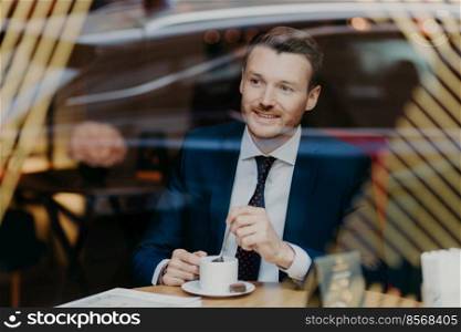 Optimistic cheerful young businessman has thoughful expression, dressed in formal clothes, looks through window in cafeteria, satisfied with results of work. People, leisure and business concept